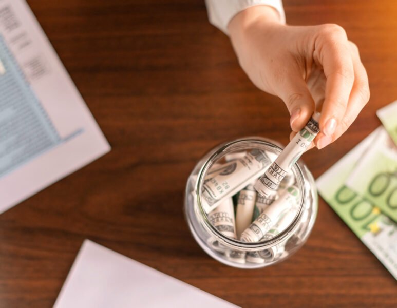 Woman dropping a banknotes into a jar with rolled banknotes on the table. Papers, money on the table