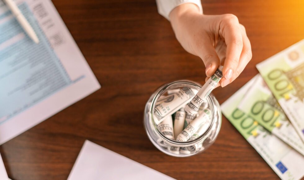 Woman dropping a banknotes into a jar with rolled banknotes on the table. Papers, money on the table